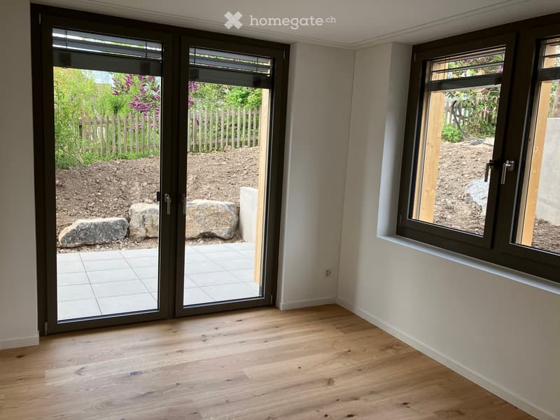 Empty room with wooden flooring, large sliding glass doors leading to a garden area, and windows showing the outdoor space.