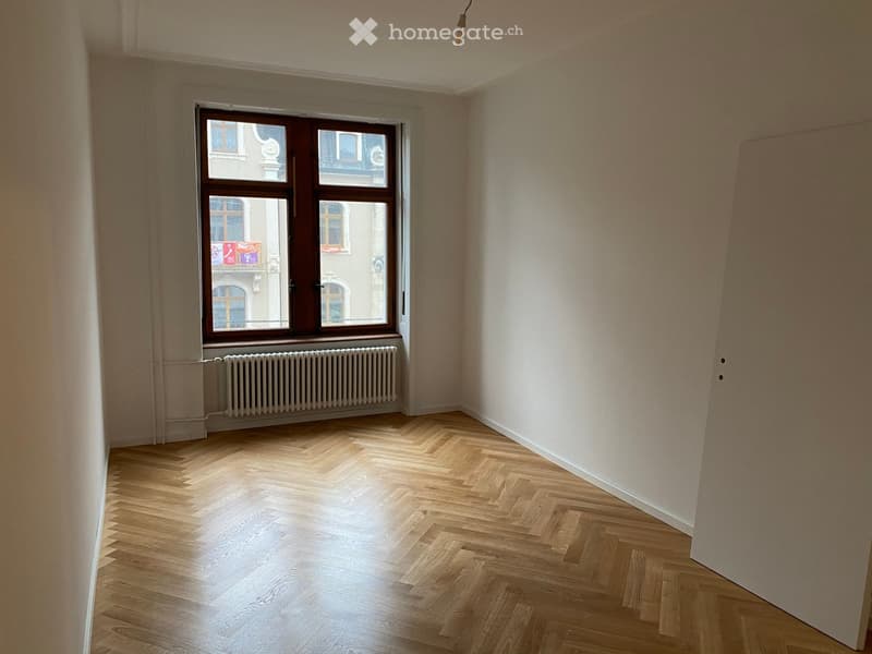 Empty room with light wooden herringbone flooring, white walls, large window with wooden frame, radiator below the window, and visible light fixture on the ceiling.