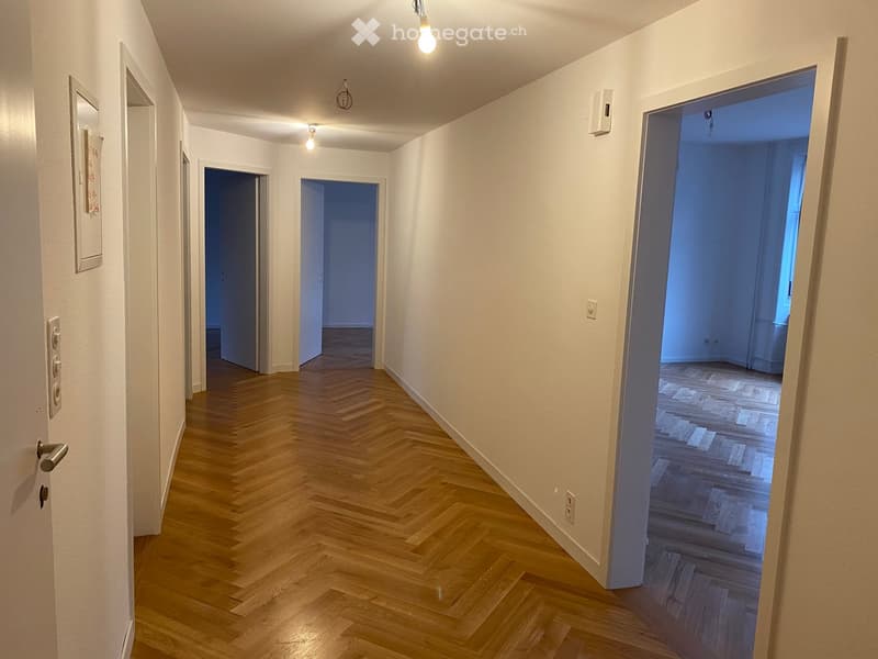 Empty hallway with wooden herringbone flooring, white walls, and several doors leading to other rooms.