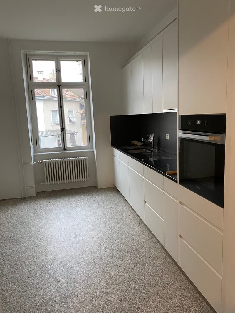 Modern kitchen with white cabinets, black countertop, sink, faucet, and built-in oven. Grey carpeted floor and large window with view of neighboring building.