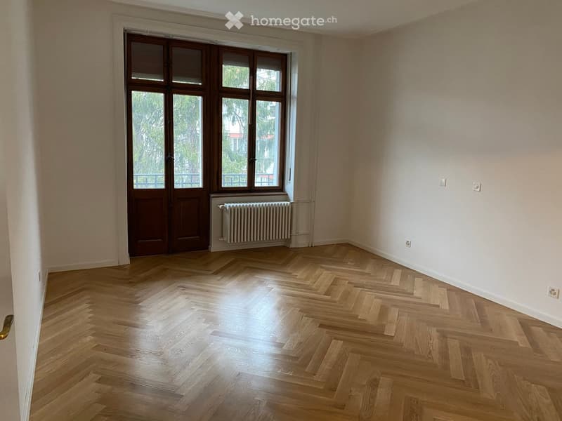 Empty room with wooden herringbone flooring, large window with double doors, radiator, and light-colored walls.