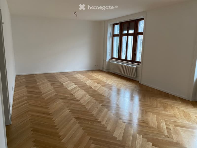 Empty room with herringbone-patterned wooden flooring, large windows with wooden frames, and a radiator beneath the windows.
