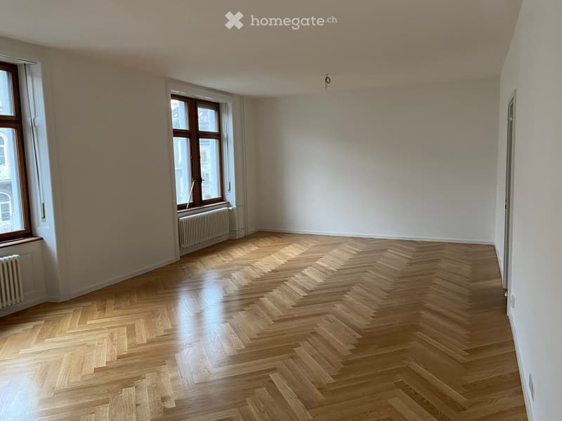 Empty room with wooden herringbone flooring, white walls, large windows, and radiators.