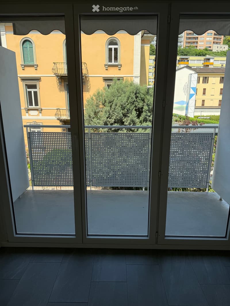 Balcony with large glass doors, white tiled floor, metal railing, and view of surrounding buildings and greenery.