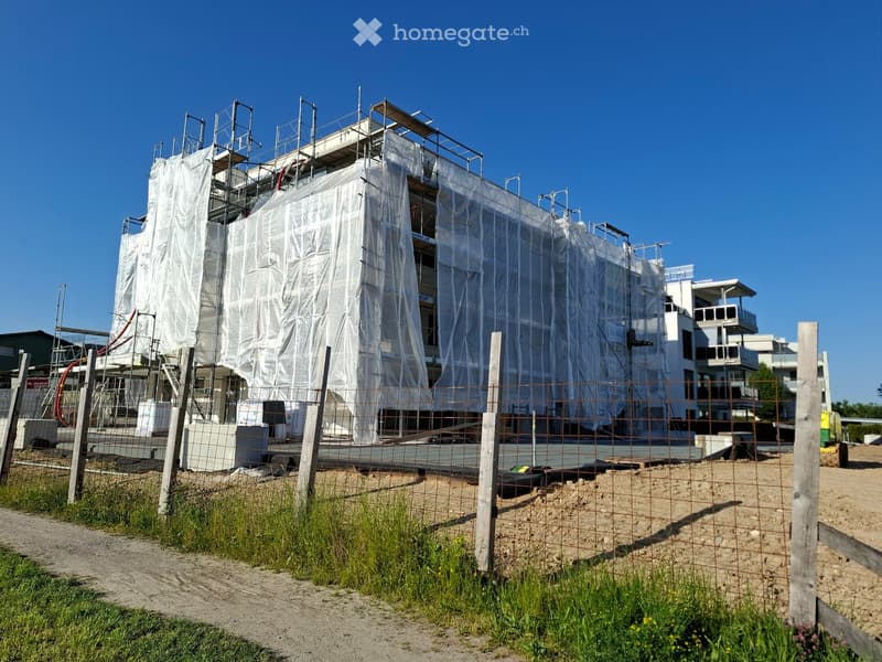 A multi-story building under construction, partially covered with scaffolding and protective sheeting, surrounded by a fenced-off construction site with exposed soil and construction materials.