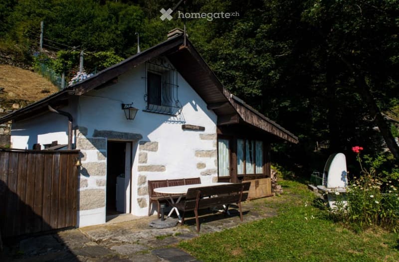 A small, rustic white stone house with a wooden roof and a stone chimney, surrounded by greenery and a wooden fence. A small outdoor seating area with a table and benches is visible in front of the house.