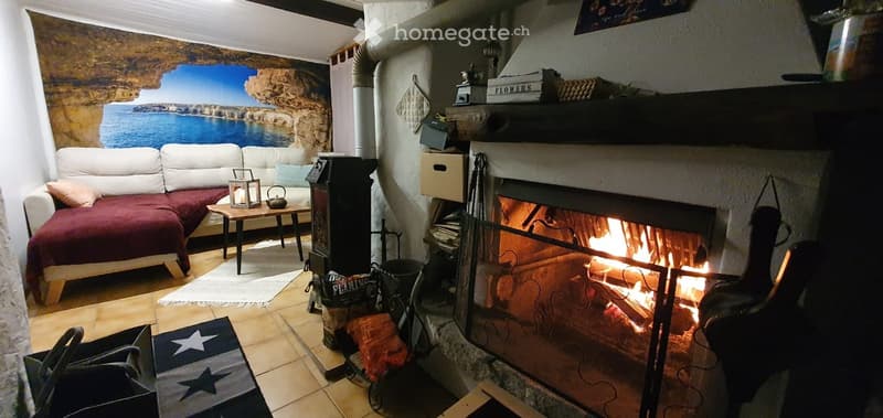 Cozy living room with a fireplace, sofa, coffee table, and decorative items. The room features a scenic mural on one wall and a warm, inviting atmosphere.