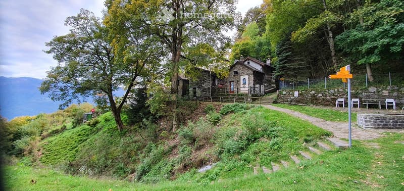 A scenic outdoor area with a stone house, grassy lawn, trees, and a pathway leading to the house. The area includes a small stone structure and a few benches.