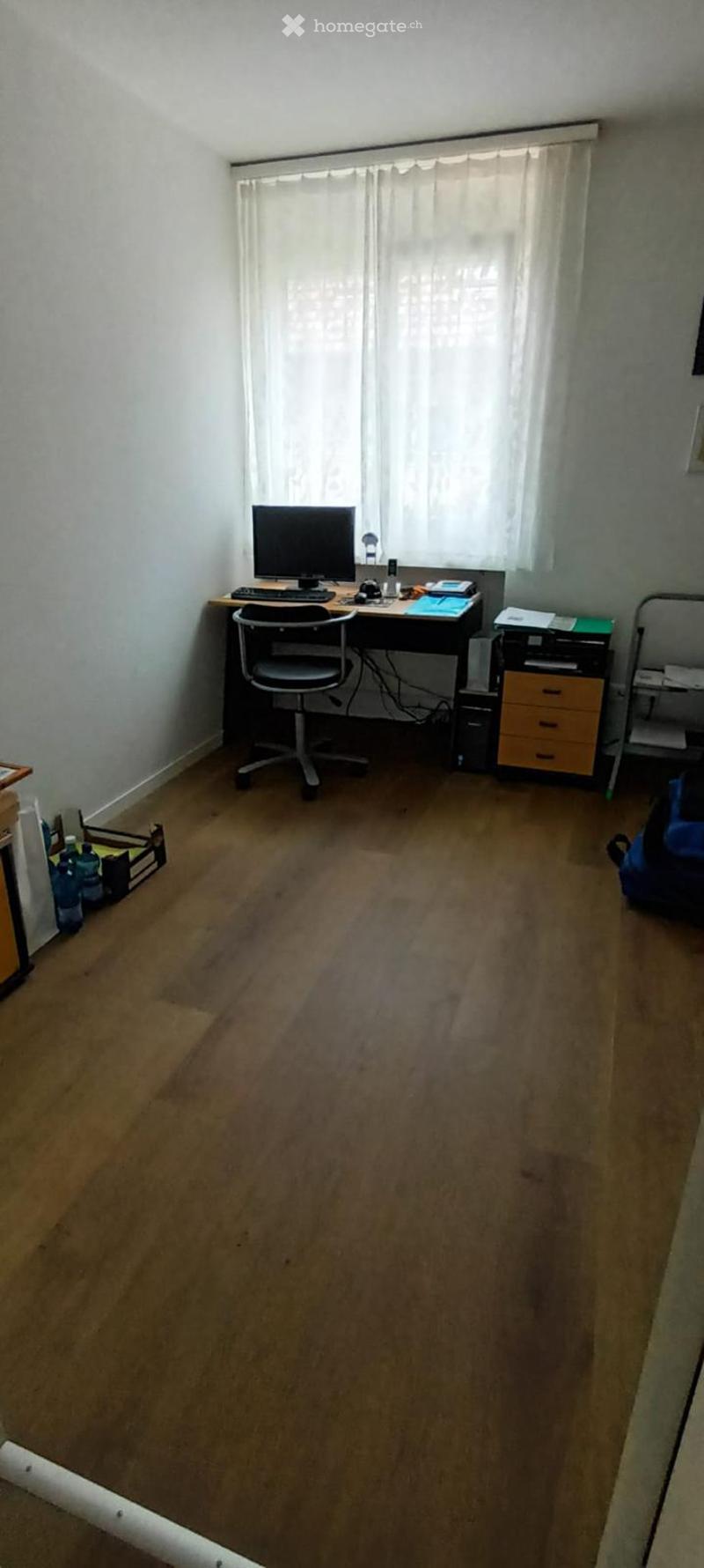 A well-lit room with wooden flooring, a desk setup including a computer, keyboard, mouse, and a chair, along with a printer and a small cabinet.