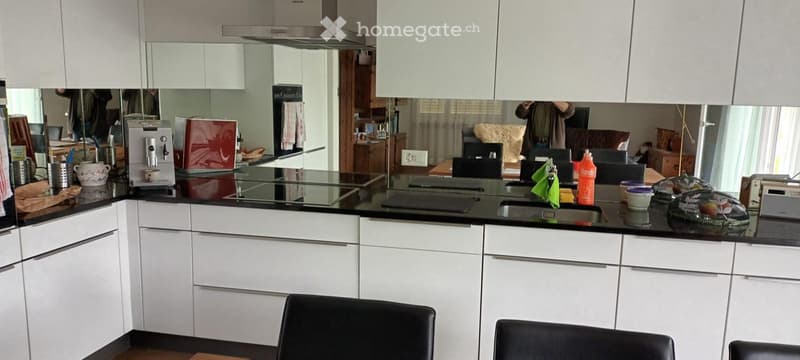 Modern kitchen with white cabinets, black countertops, and various appliances including a coffee maker and a refrigerator. There is a sink with a faucet, a bottle on the counter, and a few chairs in the foreground.