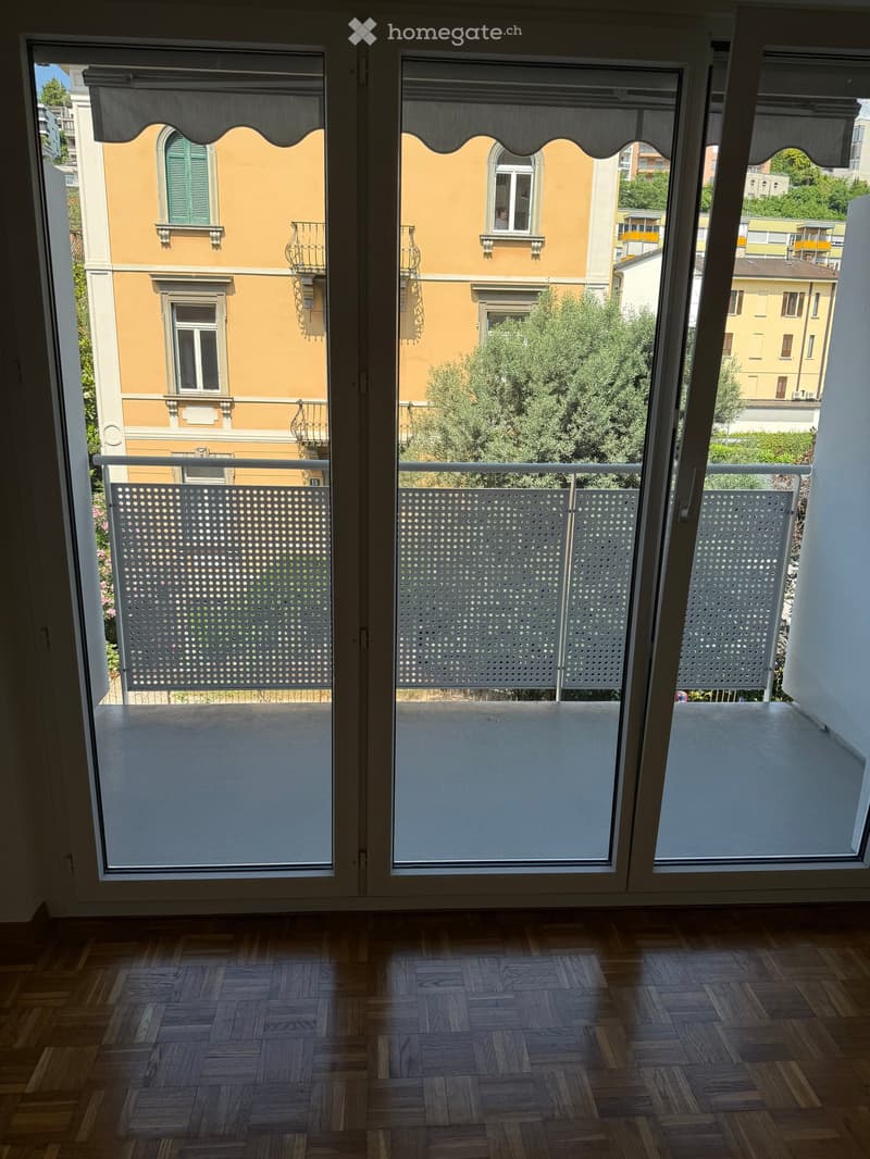 Balcony with wooden flooring, white metal railing, and a view of a yellow building with green shutters and trees.