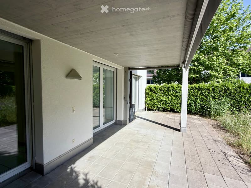Covered balcony with tiled flooring, glass sliding doors, and a view of a garden area with hedges and trees.