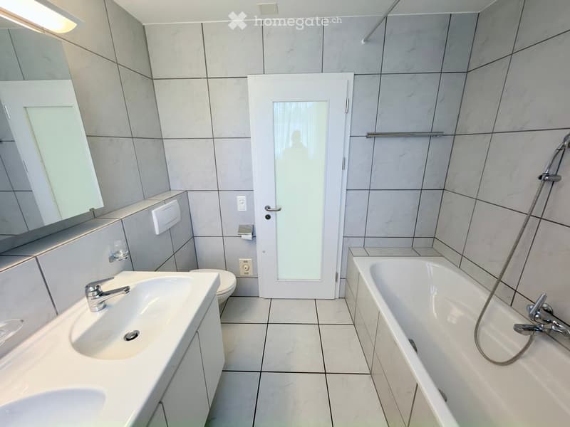 Modern bathroom with white tiles, a sink with a faucet, a bathtub, a mirror, and a toilet. Features a glass shower door and a handheld showerhead.