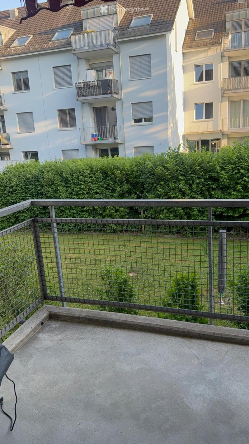 Empty rooftop terrace with metal railing, concrete surface, and view of neighboring buildings and greenery.
