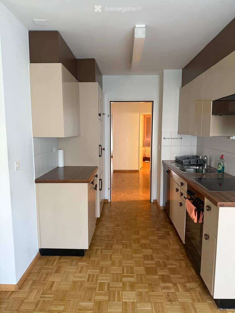Modern kitchen with light-colored cabinets, wooden herringbone flooring, and stainless steel appliances including a sink, oven, and faucet. A small hallway leads to another room.