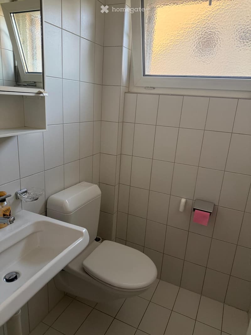 A small bathroom with a toilet, sink, and mirror. The walls and floor are tiled in light beige tiles. A window with frosted glass is visible on the right side.