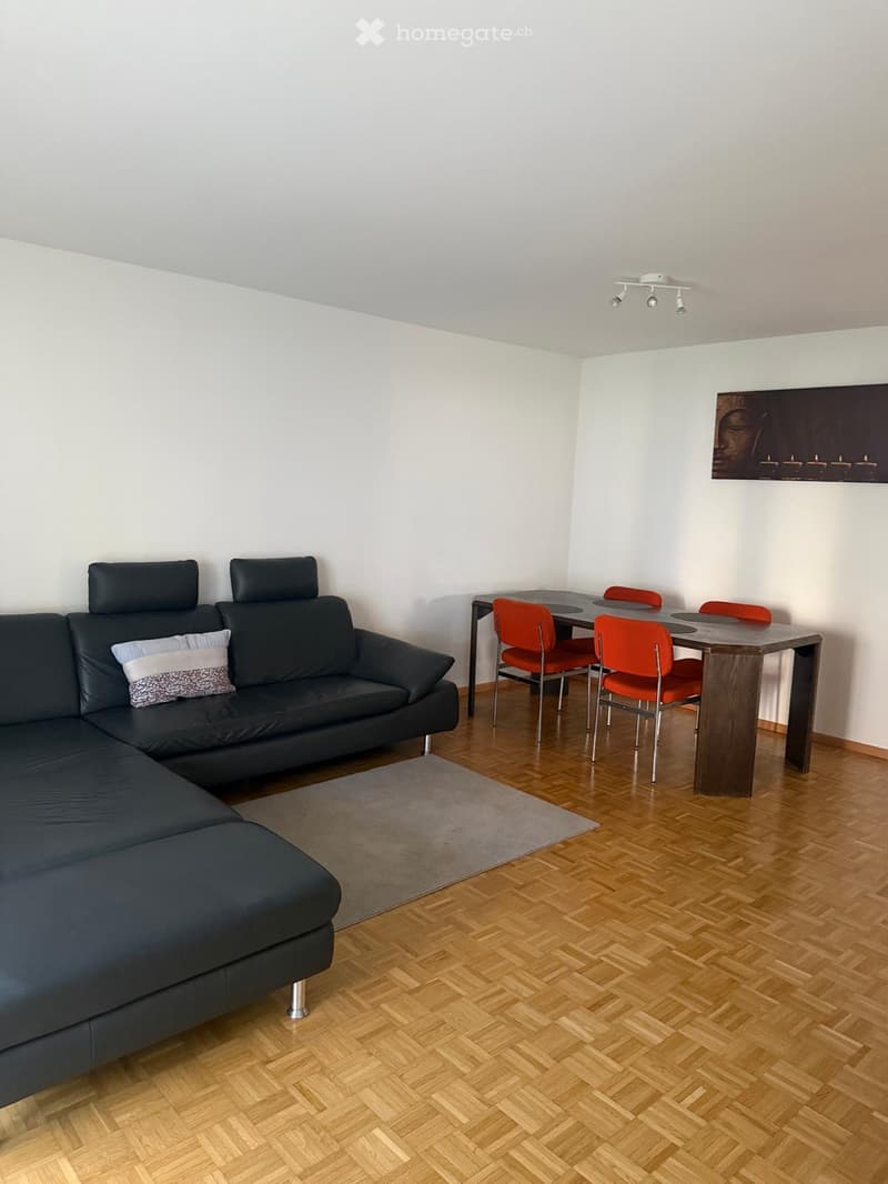 A modern living room with a dark grey sectional sofa, a wooden dining table with four orange chairs, and a grey rug on a wooden floor.