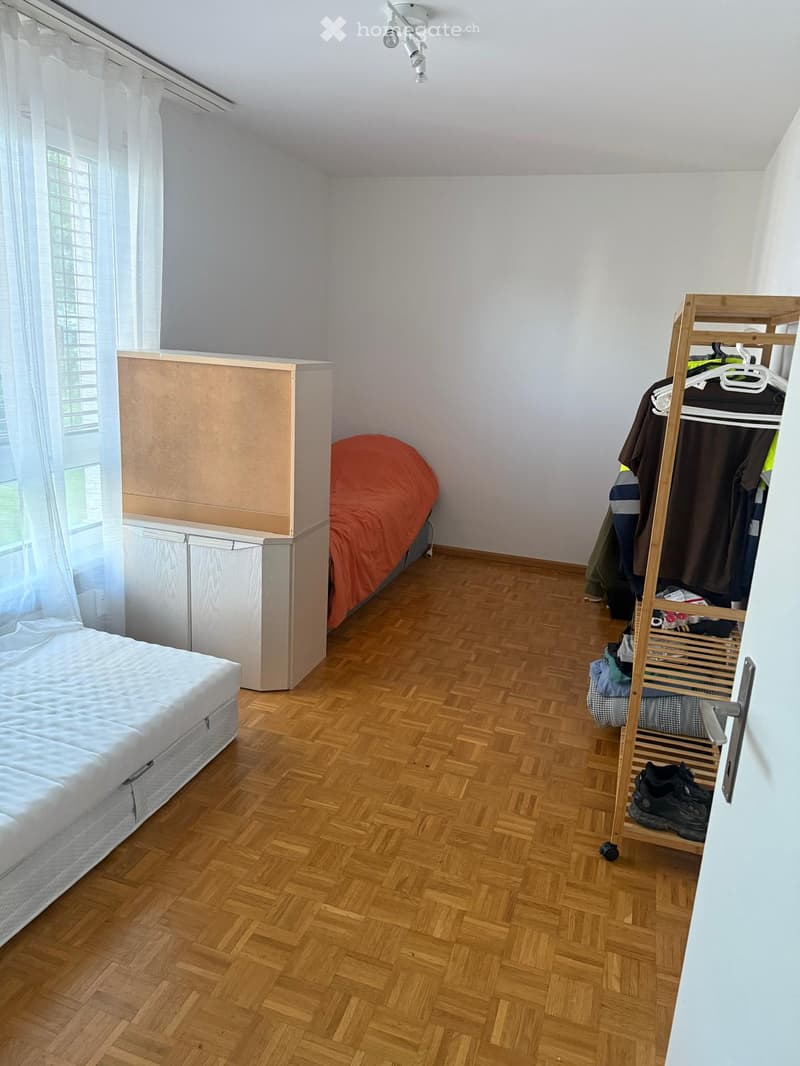 A small bedroom with wooden flooring, a single bed with white mattress, a wardrobe with clothes, and a curtained window.