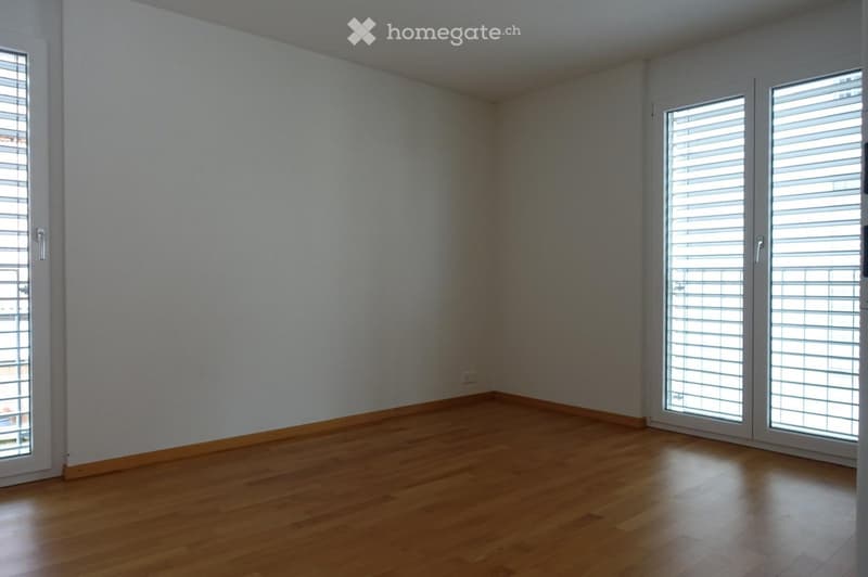 Empty room with light-colored walls and wooden flooring, featuring large windows with blinds and a door.