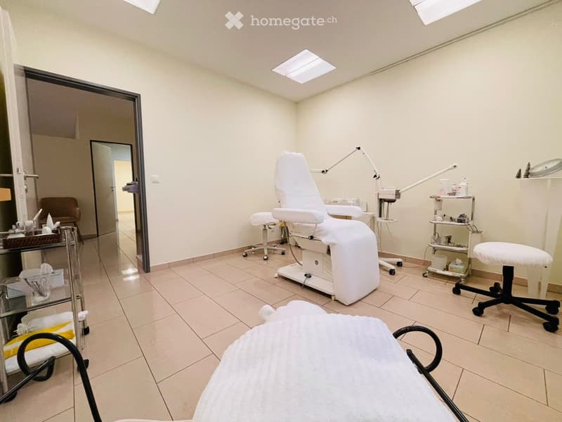 A well-lit, clean, and organized room with a white massage chair, a small white stool, a mirror, a small cabinet, and a table with various items.