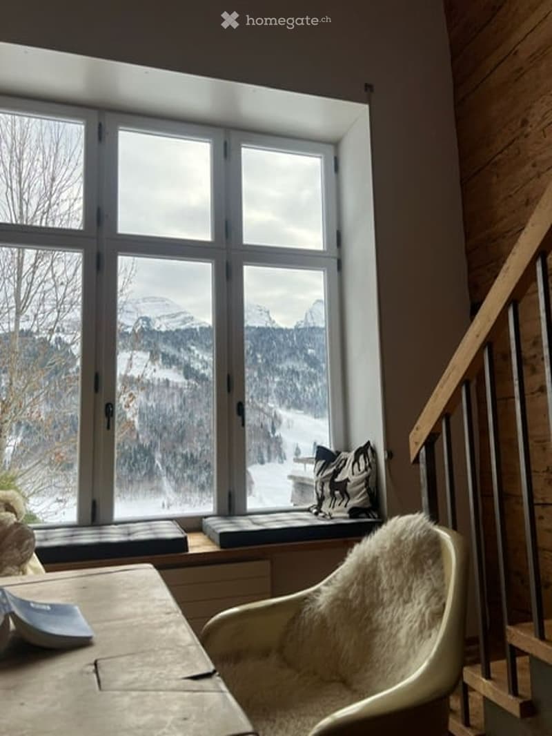 Cozy room with large window overlooking snowy landscape, featuring a fur chair, wooden table, and window bench with pillows.
