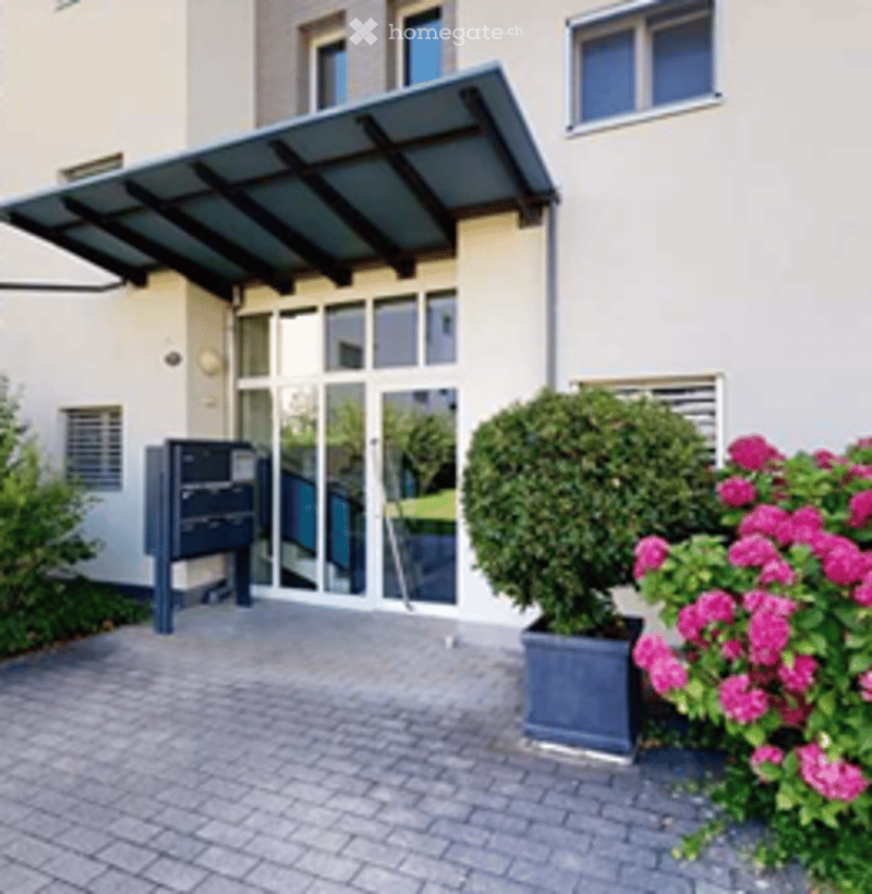 Building entrance with a covered porch, glass doors, and a mailbox. Potted plants and flowers are placed near the entrance.