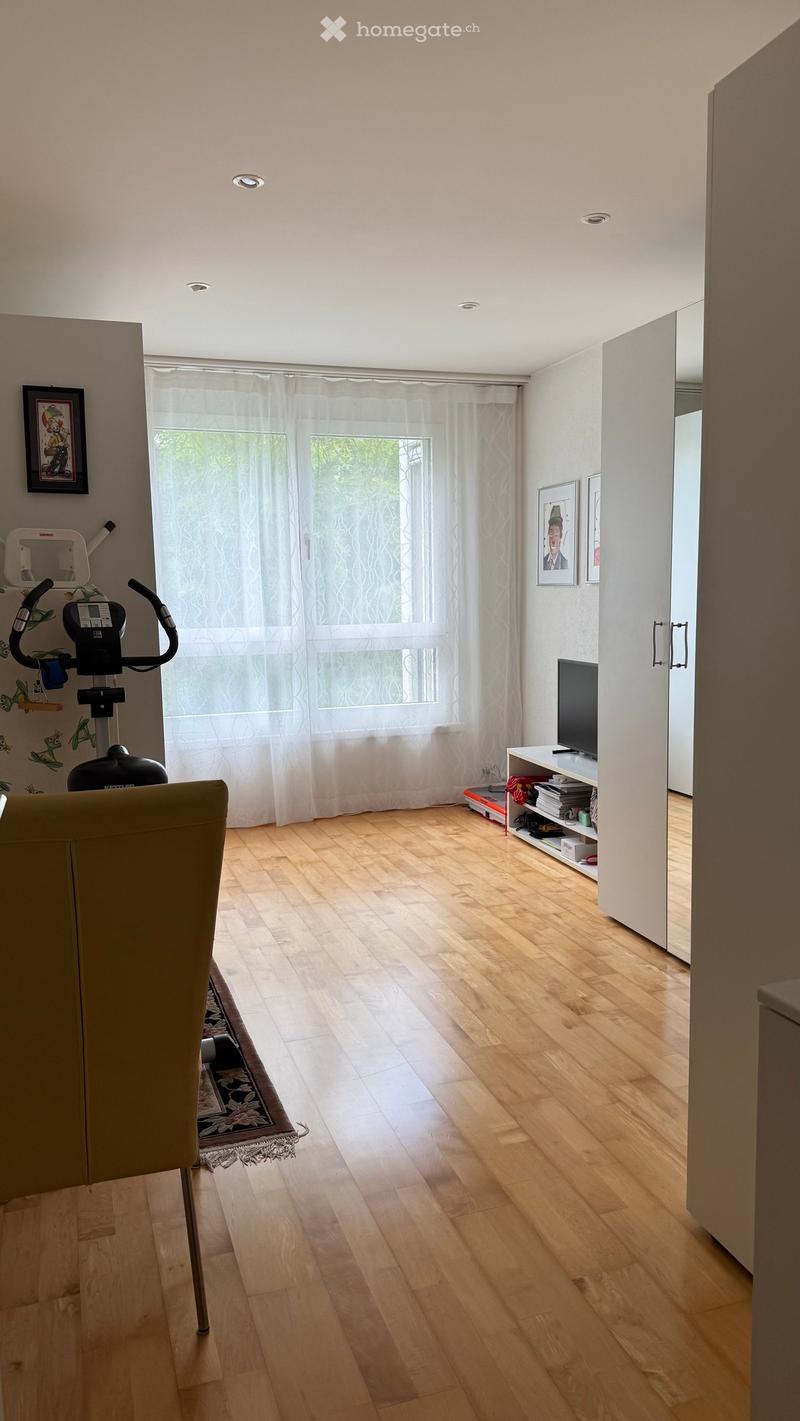 Bright living room with wooden flooring, large window with white curtains, exercise bike, TV stand, and framed pictures on the walls.