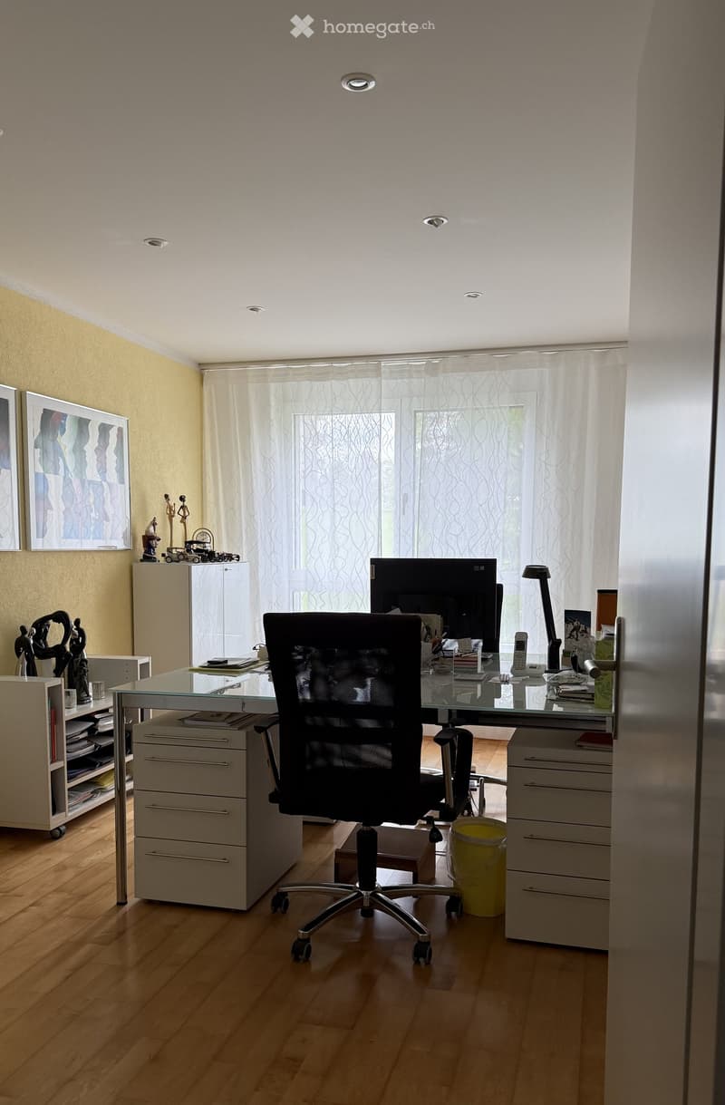 A modern office space with a glass desk, ergonomic chair, computer screen, filing cabinets, and decorative items including framed pictures and statues.