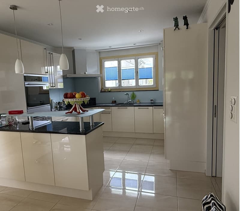 Modern kitchen with light-colored cabinets, black countertop, and a central island. Features include an oven, sink, fruit bowl, and pendant lights.