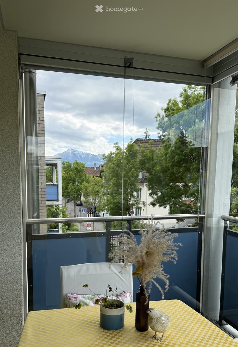 Balcony with a view of a cityscape and mountains, featuring a yellow checkered table with a vase, plant, and decorative bird figurine.