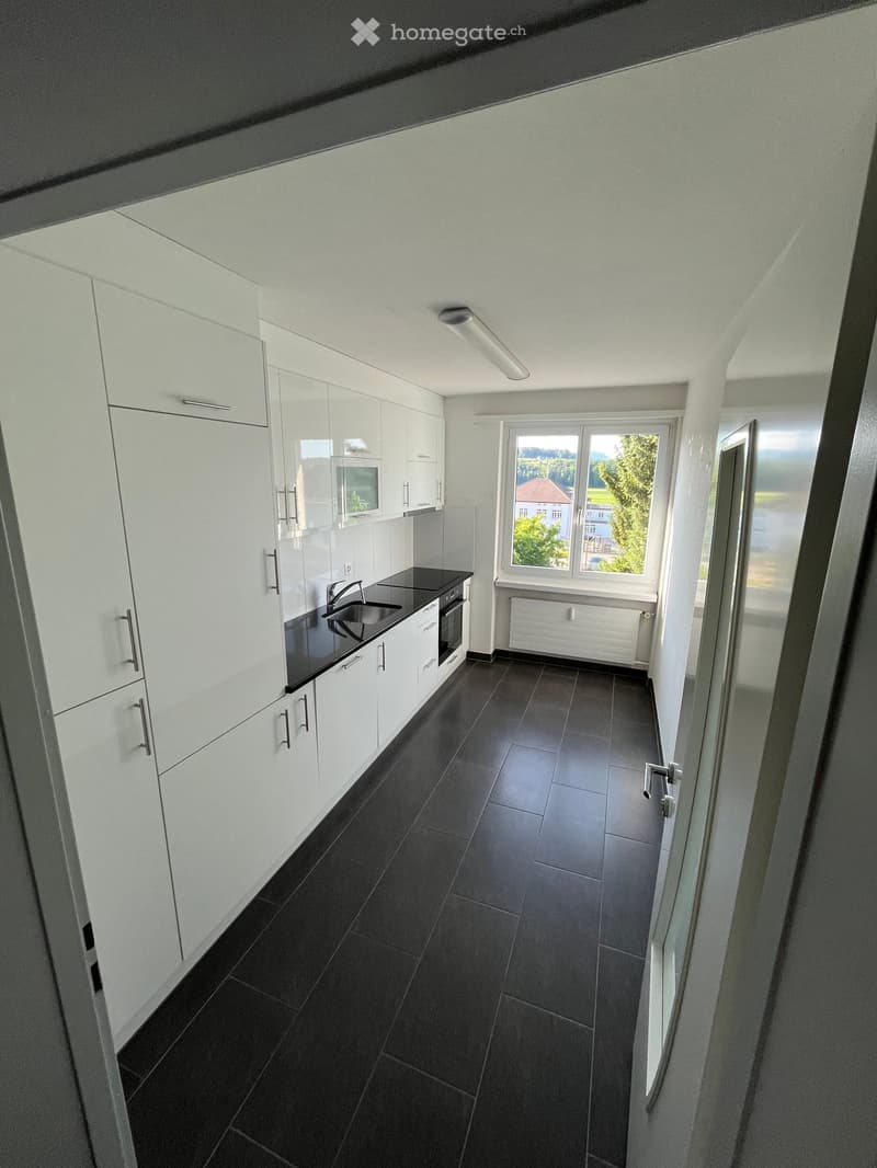 Modern kitchen with white cabinets, black countertops, and dark tiled flooring. Features a sink, faucet, and oven, with a large window offering a view of the outdoors.
