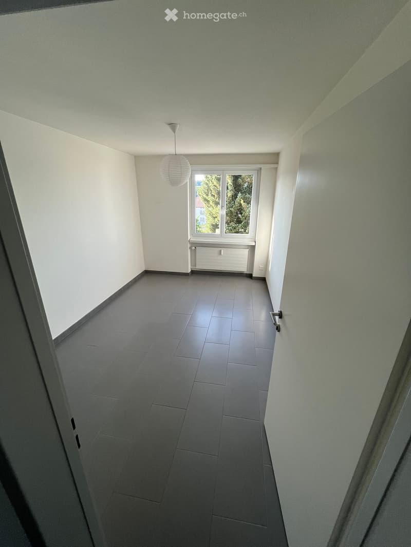 Empty hallway with grey tiled floor, white walls, a single window with a view of greenery, and a hanging spherical light fixture.