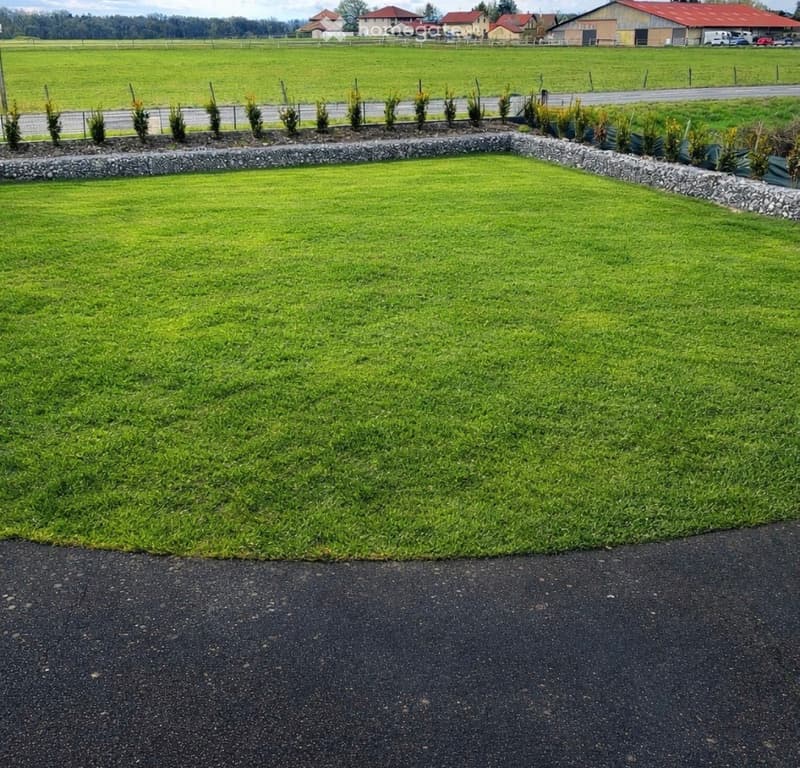 Well-maintained lawn bordered by a stone wall, with small trees and shrubs along the edge, surrounded by open fields and distant houses.