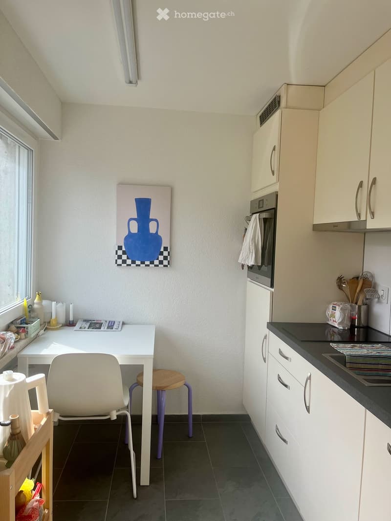 A compact kitchen with white cabinets, a black countertop, a sink, and an oven. A small white table with a chair and a stool is positioned near a window, with a painting on the wall and various small items on the table and counter.