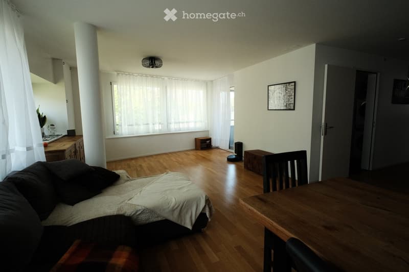 A well-lit living room with wooden flooring, a sofa, a dining table with chairs, a cabinet, a lamp, and a plant near the window.