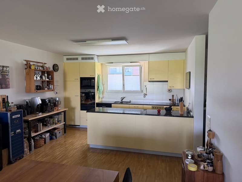 Modern kitchen with light yellow cabinets, black countertops, and various appliances including a coffee maker, blender, and microwave. Wooden shelves hold kitchen items and a small table is visible.