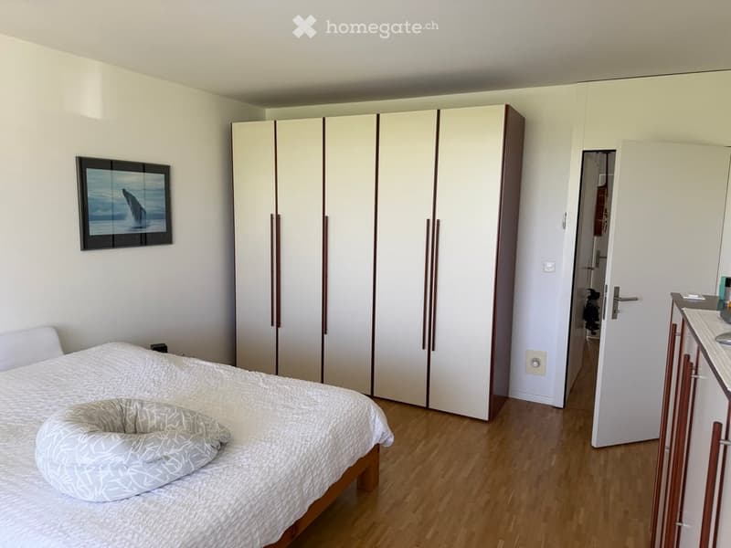 Bright bedroom with wooden flooring, a large white wardrobe, a neatly made bed with a patterned duvet, and a framed picture on the wall.