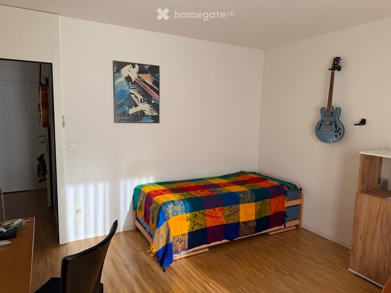 A small bedroom with a single bed covered in a colorful patchwork quilt, a wooden desk with a chair, a guitar mounted on the wall, and a picture frame above the bed.