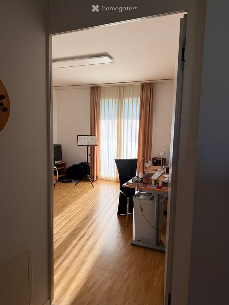 Brightly lit room with wooden flooring, a desk with a chair, a lamp, a laptop, a screen, and a tripod with a screen. There are curtains on the window and a small table with a screen in the background.