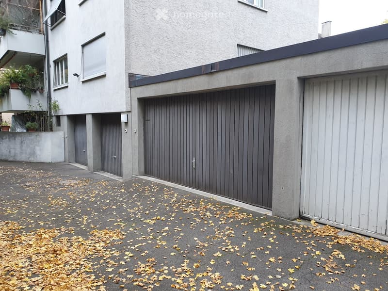 Modern multi-story building with grey garage doors, white facade, and a small balcony with potted plants.