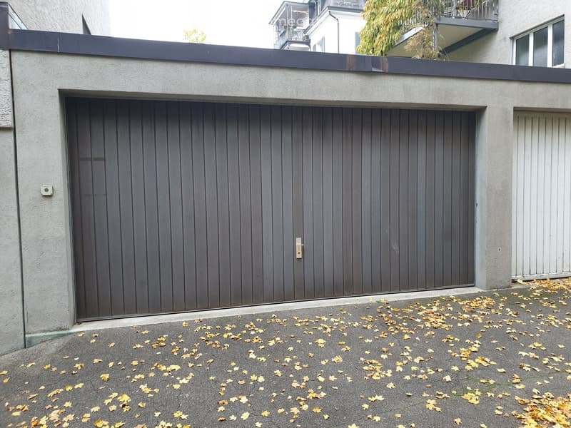 A modern building with a large, dark grey garage door and a flat roof with a dark railing. The ground is covered with fallen leaves.