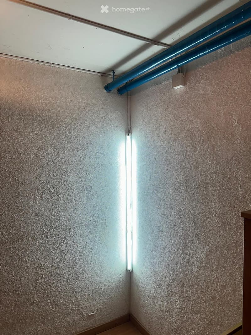 A narrow hallway with textured beige walls, a white ceiling with visible pipes, and a single fluorescent light fixture mounted on the wall.