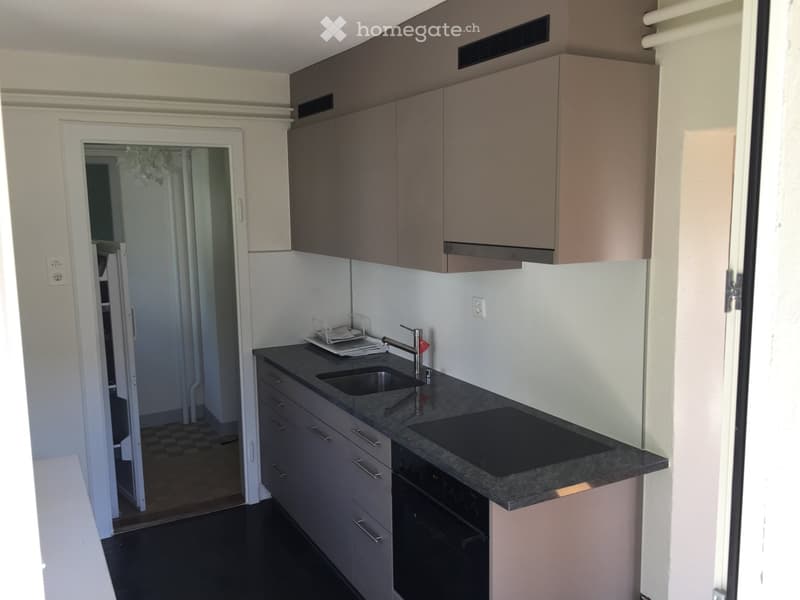 Modern kitchen with grey cabinets, black countertop, sink, faucet, and built-in oven. Open doorway leads to another room.