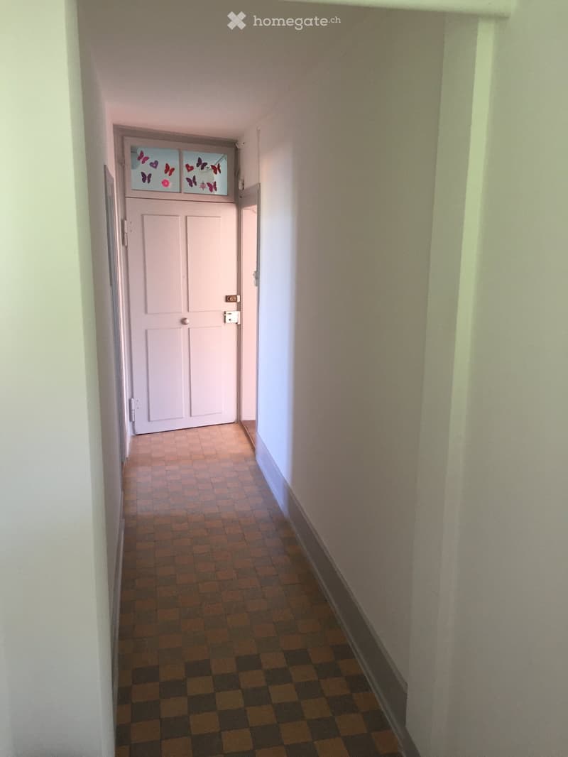 A narrow hallway with beige walls, a tiled floor, and a white door at the end featuring a decorative glass panel with butterfly designs.