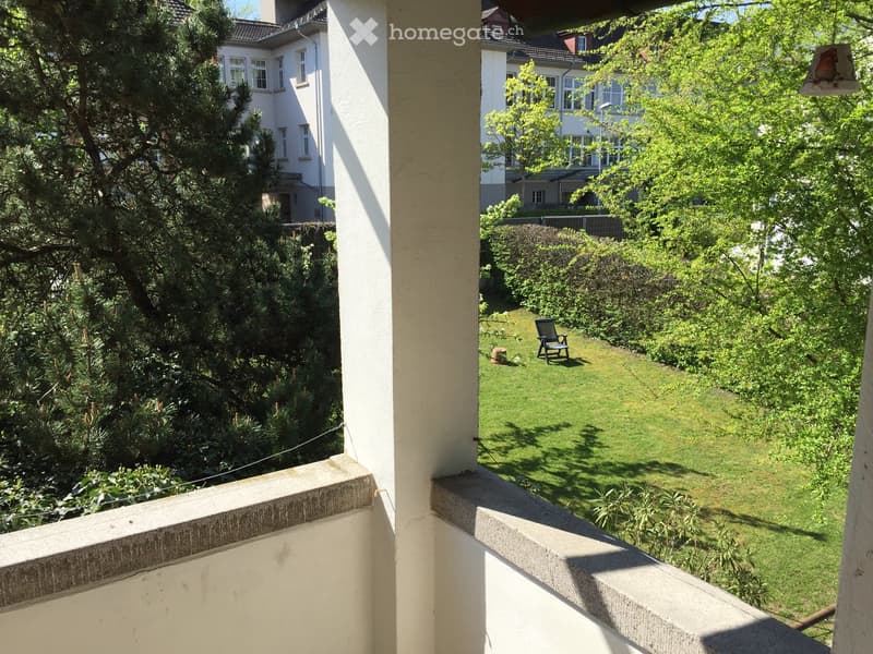 Balcony overlooking a well-maintained garden with green grass, trees, and a single chair placed on the lawn.