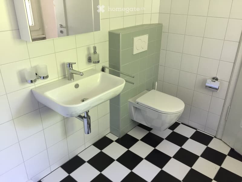 Modern bathroom with white tiled walls, black and white checkered floor, white sink with chrome faucet, toilet, mirror, and soap dispenser.