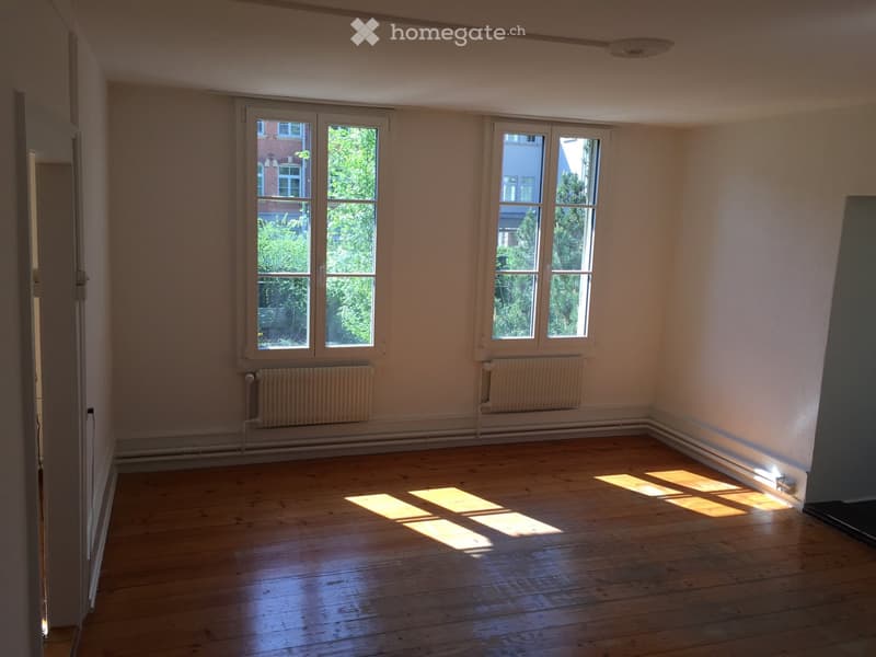 Empty room with wooden flooring, large windows, radiators, and natural light streaming in.