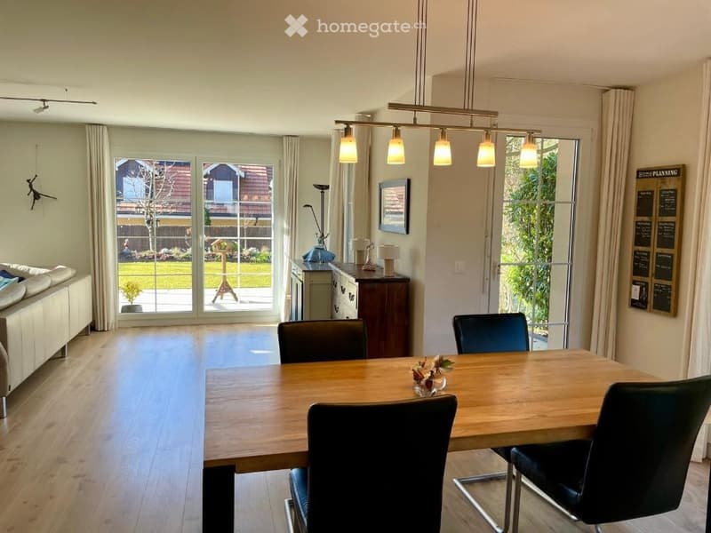 Bright, open-plan living and dining area with wooden flooring, large windows, a sofa, a dining table with chairs, and a cabinet with decorative items.