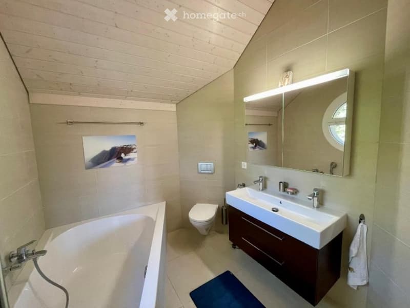 Modern bathroom with a bathtub, toilet, double sink, large mirror with built-in lighting, and a dark wooden cabinet.