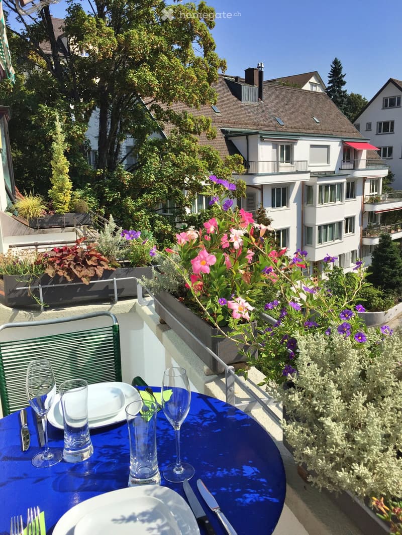 Outdoor dining area on a balcony with a blue table, plants, and a view of a building with a terrace and plants.